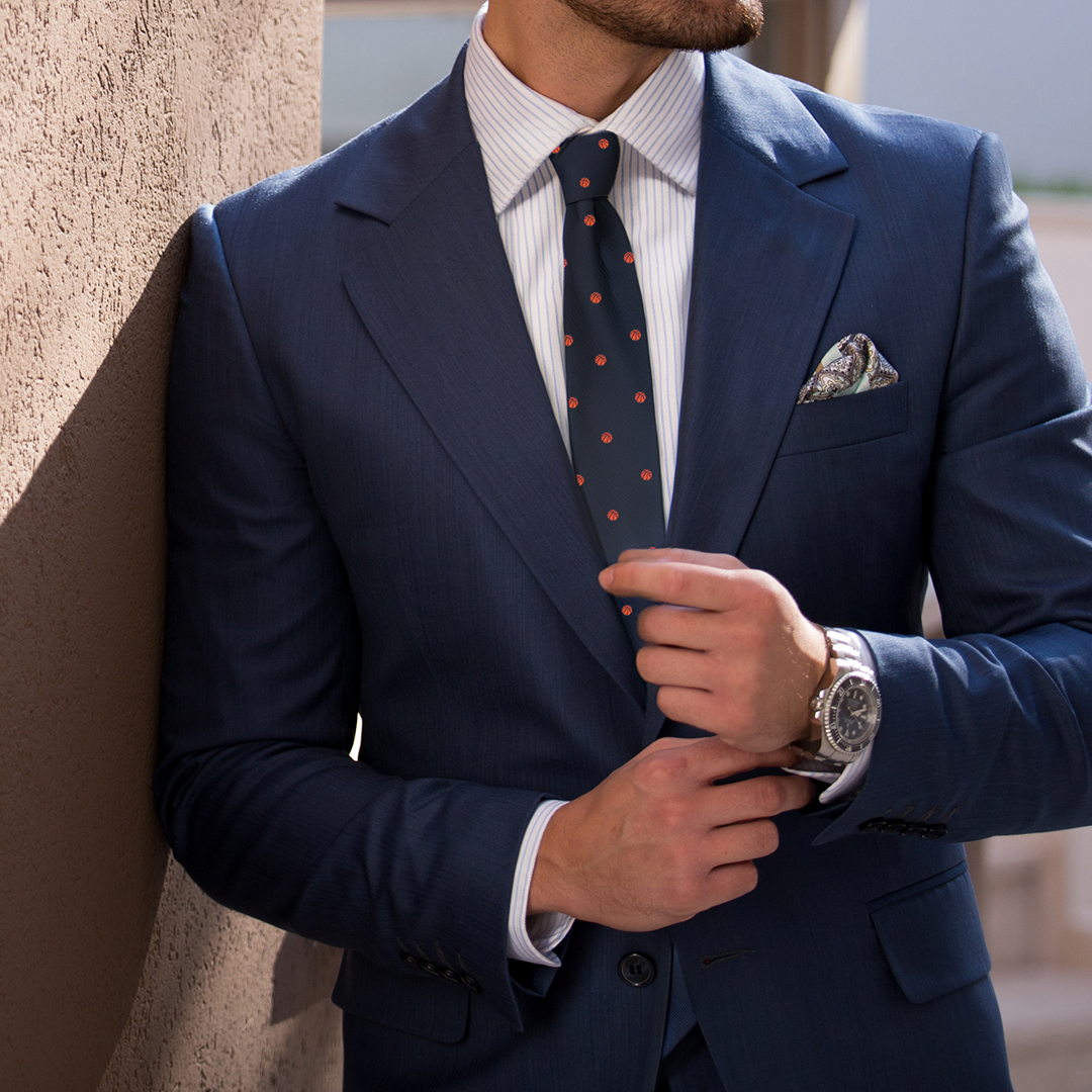 A man in a blue suit with a striped shirt and Basketball Thin Tie adjusts his wristwatch with elegance while standing against a wall.