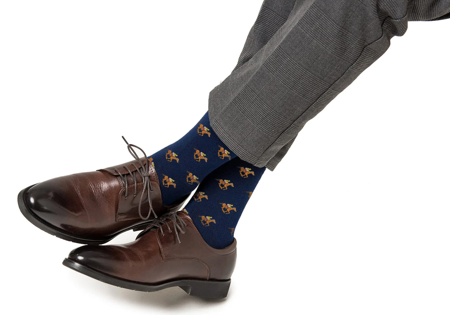 A person wearing gray plaid dress pants, brown dress shoes, and Melbourne Race Horse Socks with a horse pattern has their feet crossed against a white background.