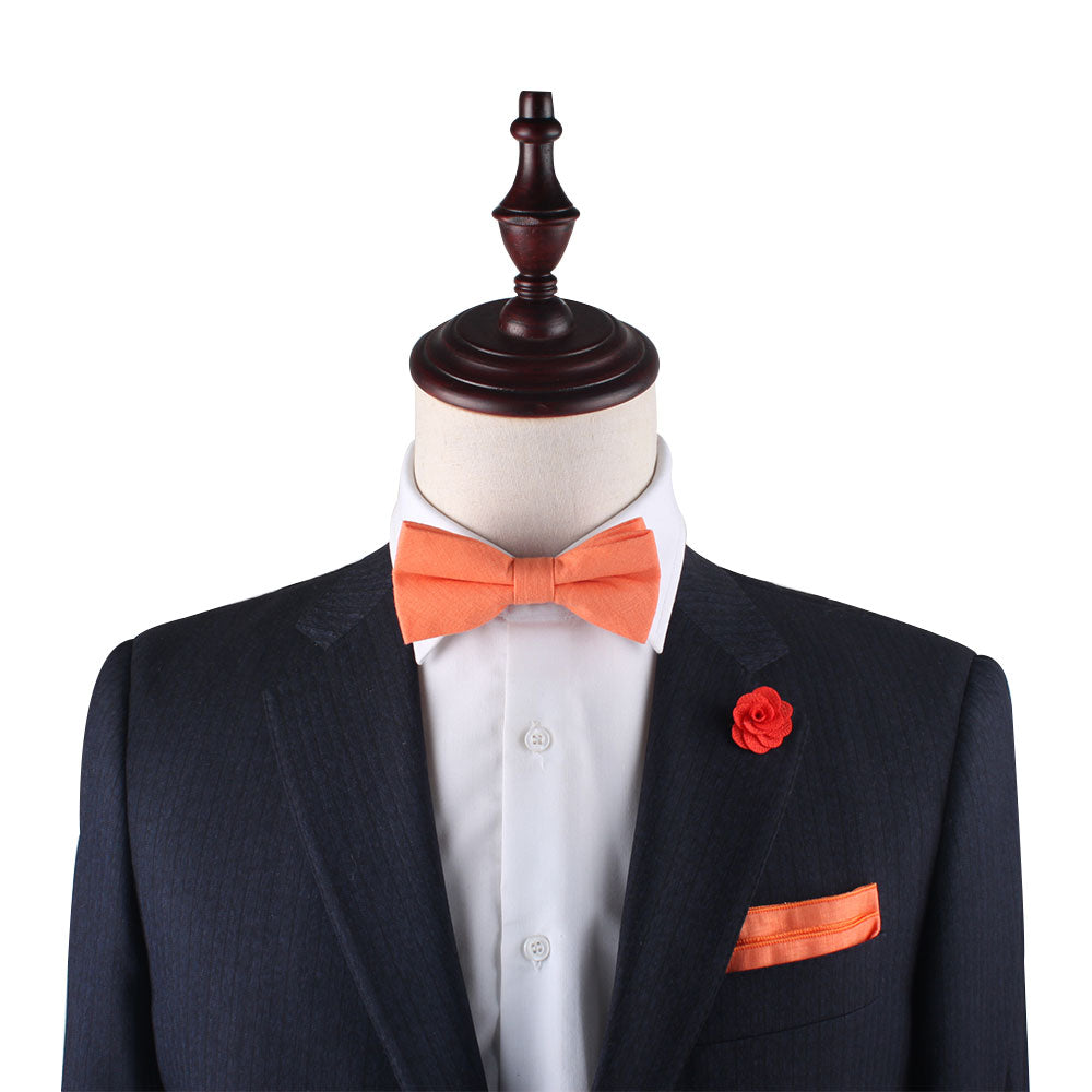 A mannequin showcases spring elegance in a dark suit and white shirt, featuring the Peach Orange Cotton Bow Tie & Pocket Square Set, accented by an orange lapel flower.