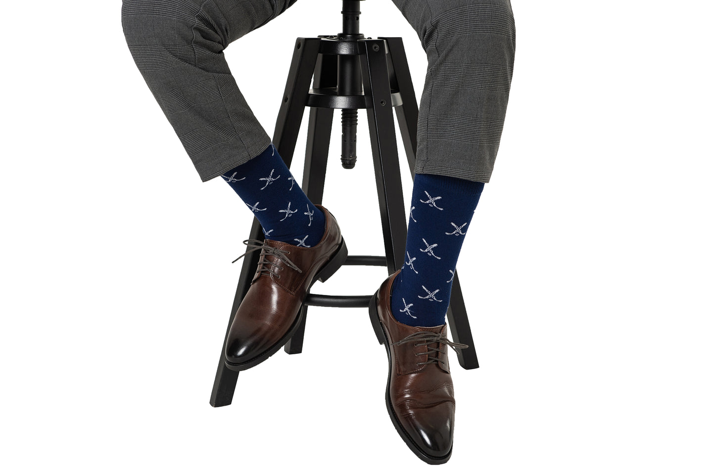 A man exudes style, sitting on a black stool in his brown shoes, gray pants, and Crossed Ice Hockey Socks.