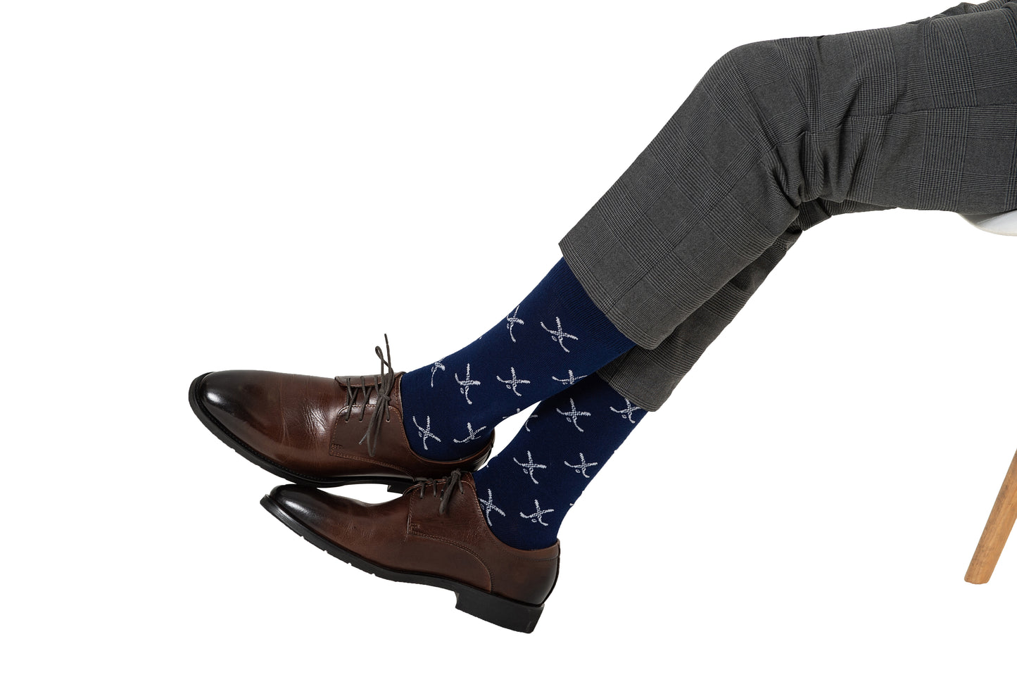 A man, showcasing his style, is sitting cross-legged in gray trousers, dark brown shoes, and blue socks adorned with a crossed ice hockey pattern.