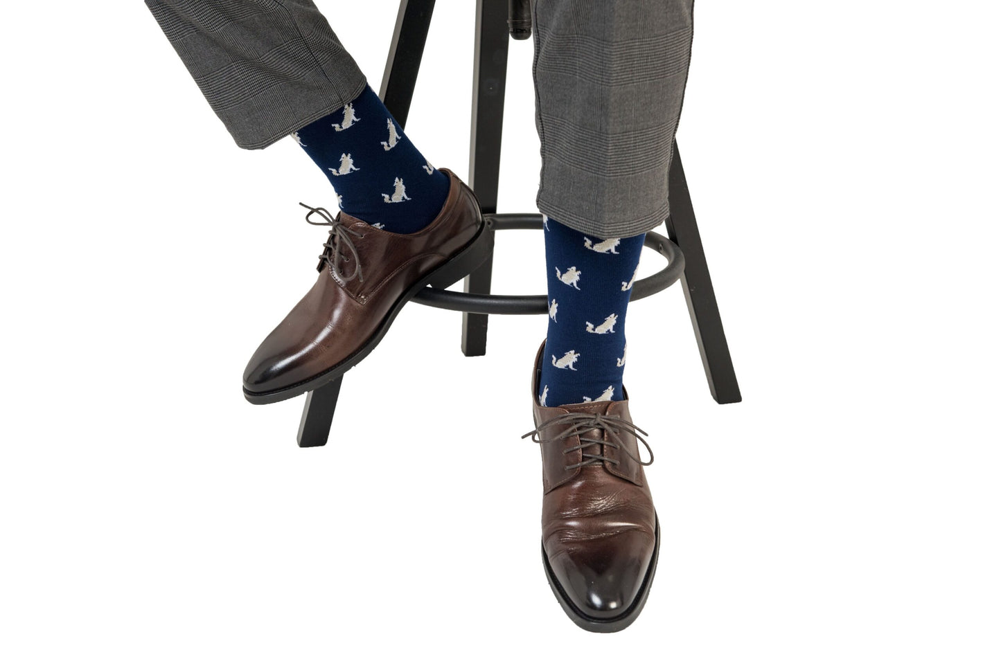A person, set to lead the pack with every step, wearing stylish brown shoes and Wolf Socks with white bird patterns seated on a black stool.