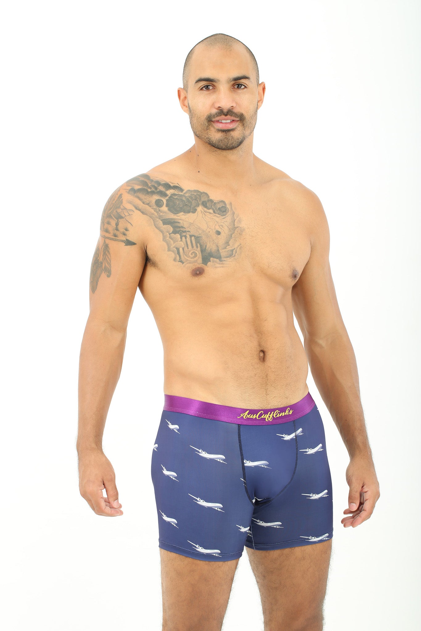 A man with a tattoo on his shoulder is modeling the relaxed style of Airplane Underwear, featuring cozy purple boxer briefs adorned with a pink waistband and shark pattern.