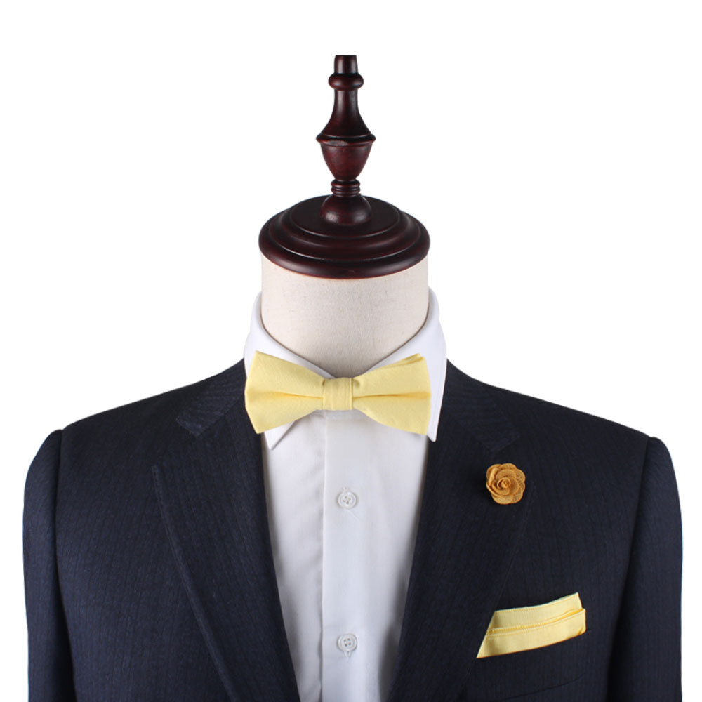 A mannequin wearing a dark suit and white shirt is highlighted with lively accents featuring a yellow bow tie, pocket square named "Baby Yellow Pocket Square," and a flower lapel pin—ideal for adding flair to any occasion.