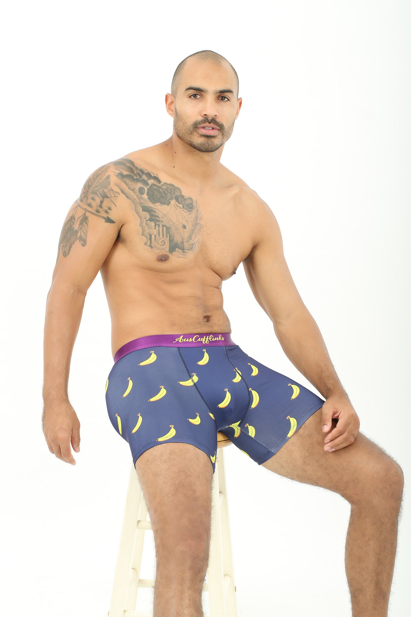 A man with a tattoo on his shoulder is sitting on a plush stool, wearing navy Banana Underwear featuring a banana peel pattern.