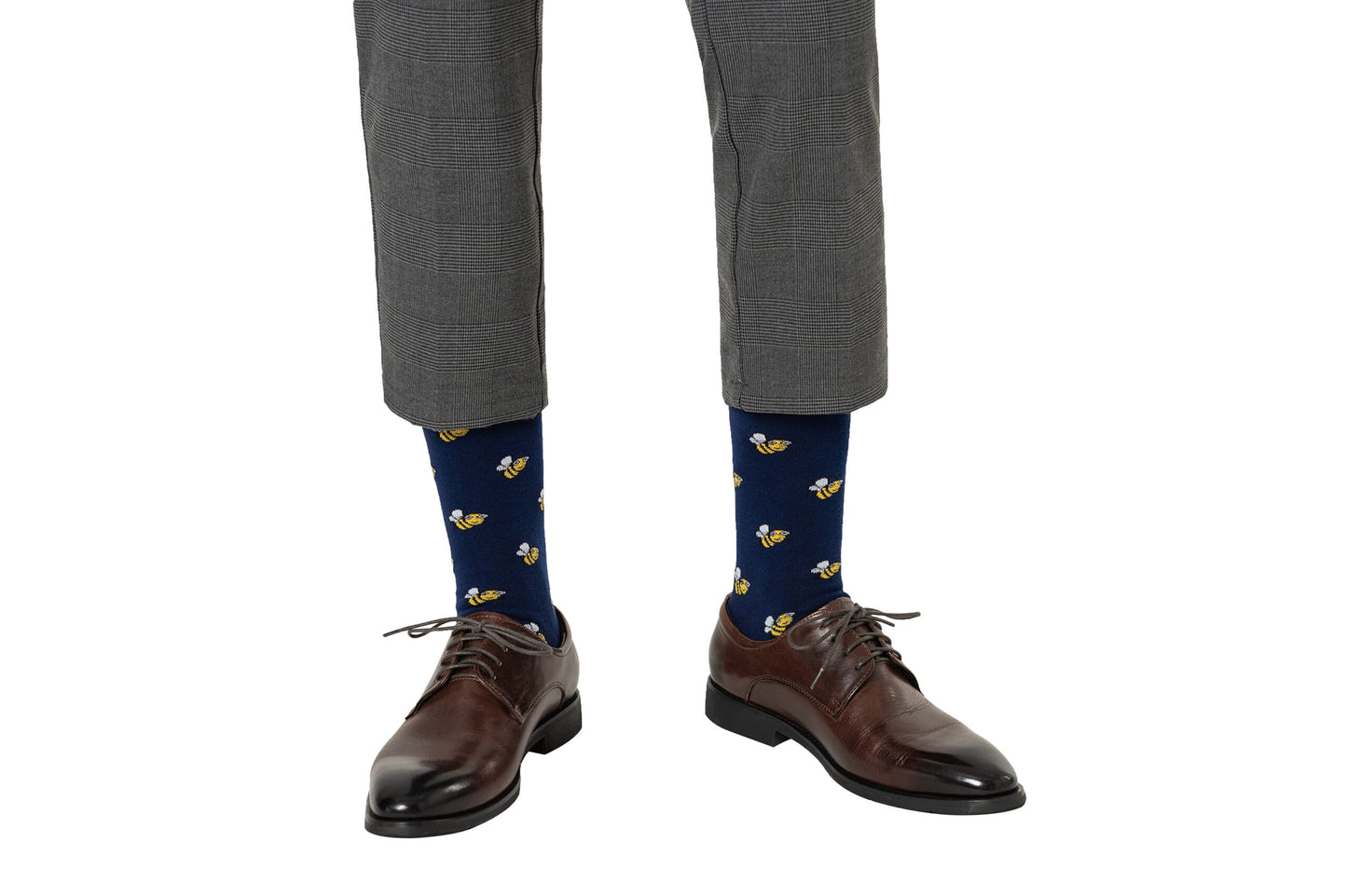 A person stands on a white background wearing gray plaid pants, brown dress shoes, and Bee Socks in navy blue with a bee print.