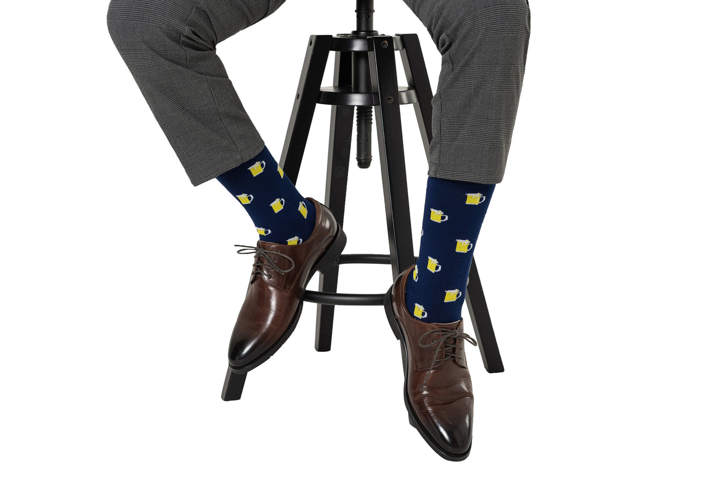 A person wearing Beer Socks—navy blue with yellow beer mug patterns—pairs them with gray dress pants and brown shoes while sitting on a black stool, making these socks perfect for beer lovers.