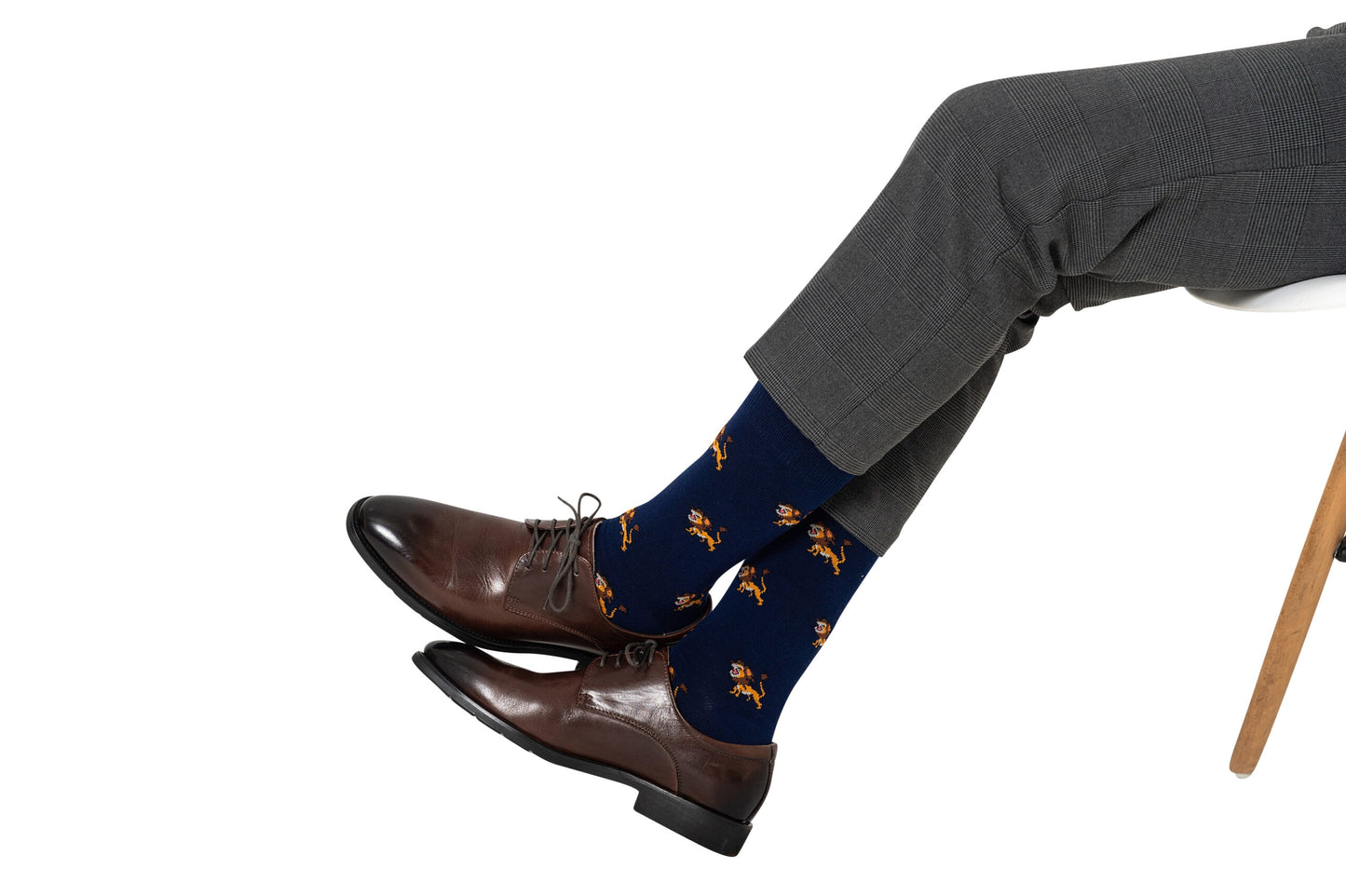 A person in gray trousers and brown dress shoes sits with legs crossed, wearing Lion Socks featuring a wildlife lion pattern, on a white background.