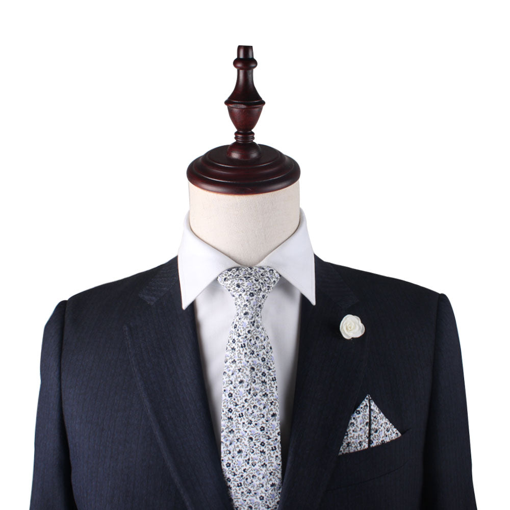 A mannequin dressed in a black suit with a white collared shirt and floral tie, complemented by the Black Print Floral Pocket Square and a white lapel pin, is given a subtle hint of lavender to enhance its style.