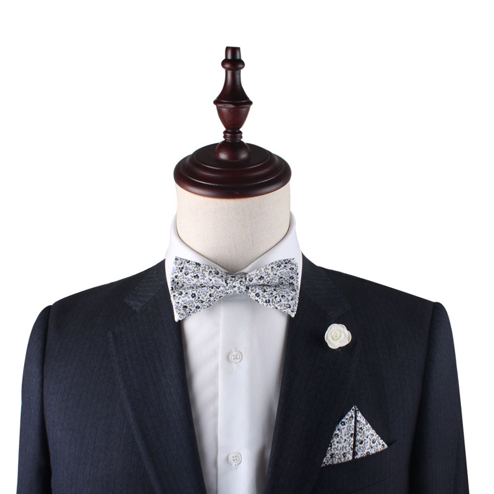 A tailor's mannequin dressed in a dark suit with a white shirt, patterned bow tie, and a Black Print Floral Pocket Square adorned with a white flower lapel pin embodies subtle style with a hint of lavender elegance.