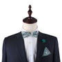 A mannequin in a dark suit displays the Teal Floral Bow Tie, complemented by a white shirt, green floral pocket square, and matching green lapel pin against a plain background—ideal for weddings or formal events.