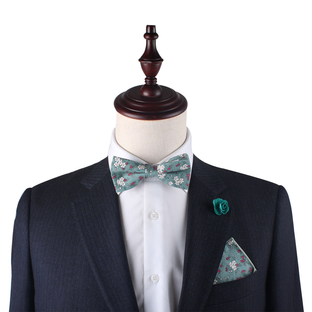 A mannequin in a dark suit displays the Teal Floral Bow Tie, complemented by a white shirt, green floral pocket square, and matching green lapel pin against a plain background—ideal for weddings or formal events.