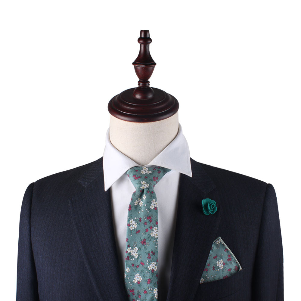 A tailored suit on a mannequin features a dark jacket, white shirt, floral tie, and the Teal Floral Pocket Square. Completing the ensemble is a green lapel flower, all beautifully arranged on a wooden stand.