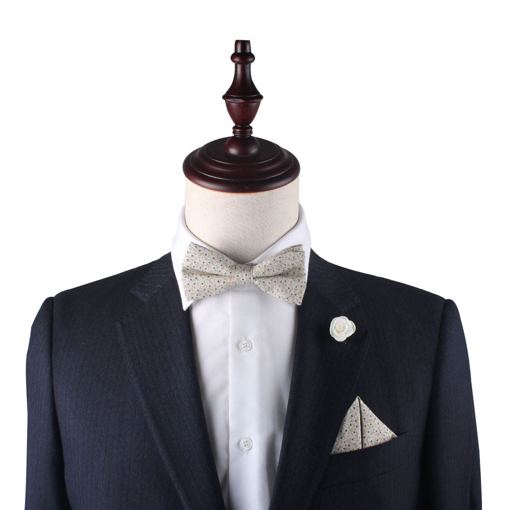 A mannequin dressed in a black suit with a white shirt, patterned bow tie, and the Blue Forget-Me-Nots Floral Pocket Square exudes subtle sophistication. A decorative white flower is pinned to the lapel, completing this statement of class.
