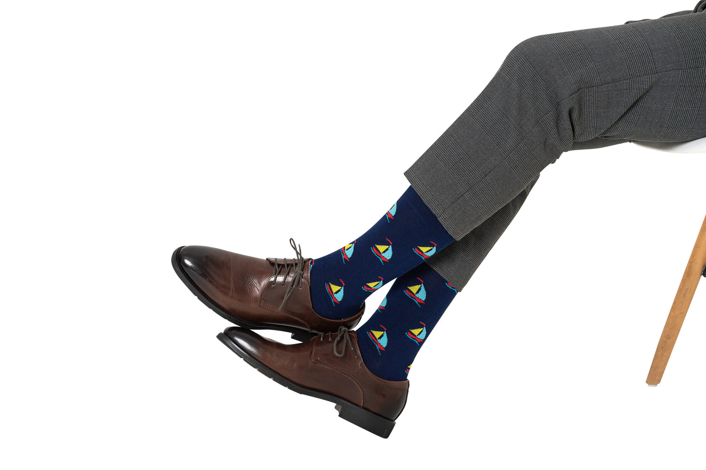 Wearing grey dress pants, brown leather shoes, and vibrant Sail Boat Socks featuring colorful sailboat patterns—ideal for ocean lovers—a person sits cross-legged on a wooden chair.