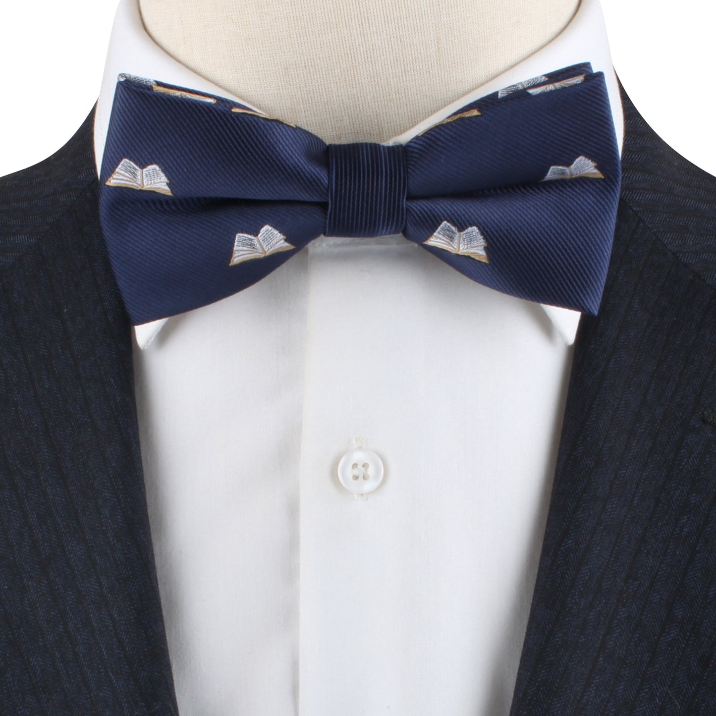 A close-up view of the Book Bow Tie, showcasing its blue fabric adorned with an open book pattern, worn elegantly with a white dress shirt and a dark pinstriped suit, perfectly scripting your style story.