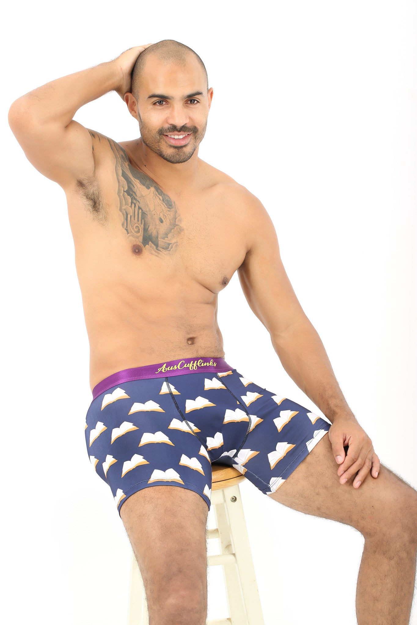 A shirtless man with a chest tattoo sits on a stool, wearing Book Underwear – Fun Gift for Book Lovers featuring blue patterns and a purple waistband—ideal for those passionate about literature.