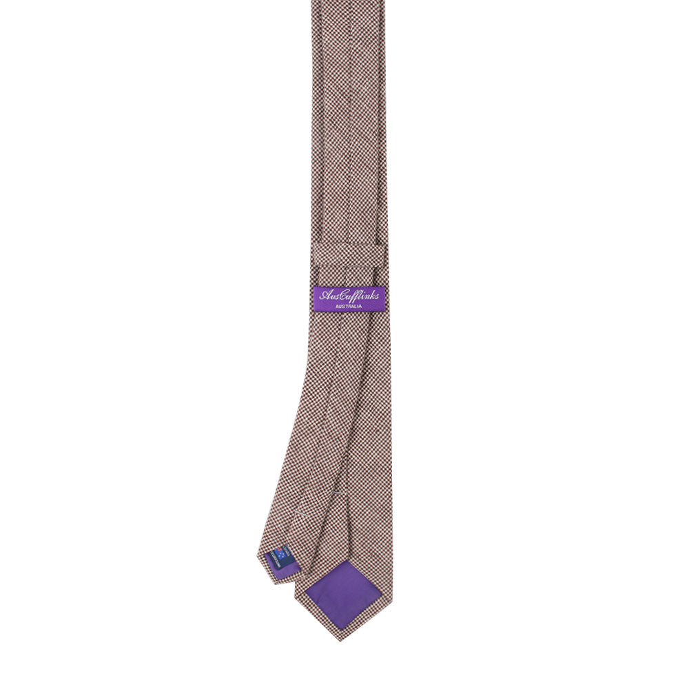 The Brown Mini Houndstooth Cotton Skinny Tie exudes elegance with its pattern, a purple label on the back, and a slender strip of purple fabric underneath.