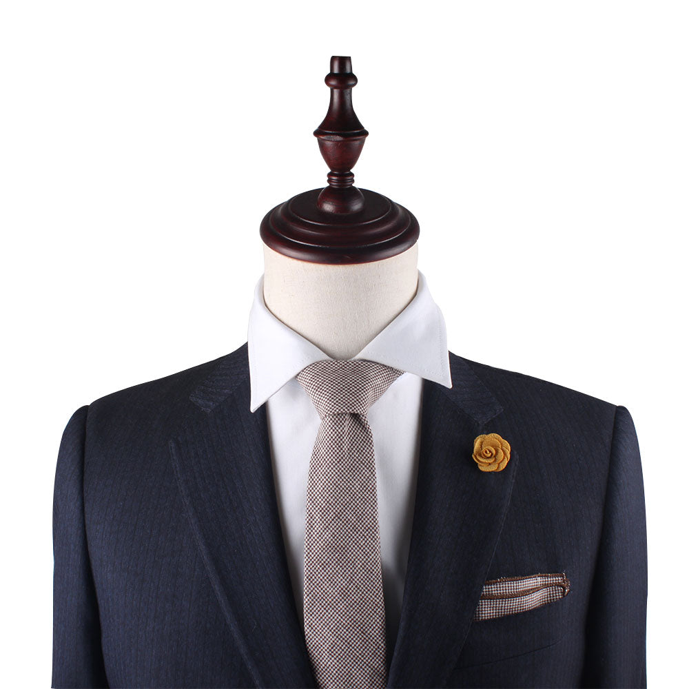 A mannequin, dressed in a dark suit with a white shirt, beige tie, and yellow lapel flower against a plain backdrop, highlights sophistication. This ensemble is perfectly accented by the Brown Mini Houndstooth Pocket Square.
