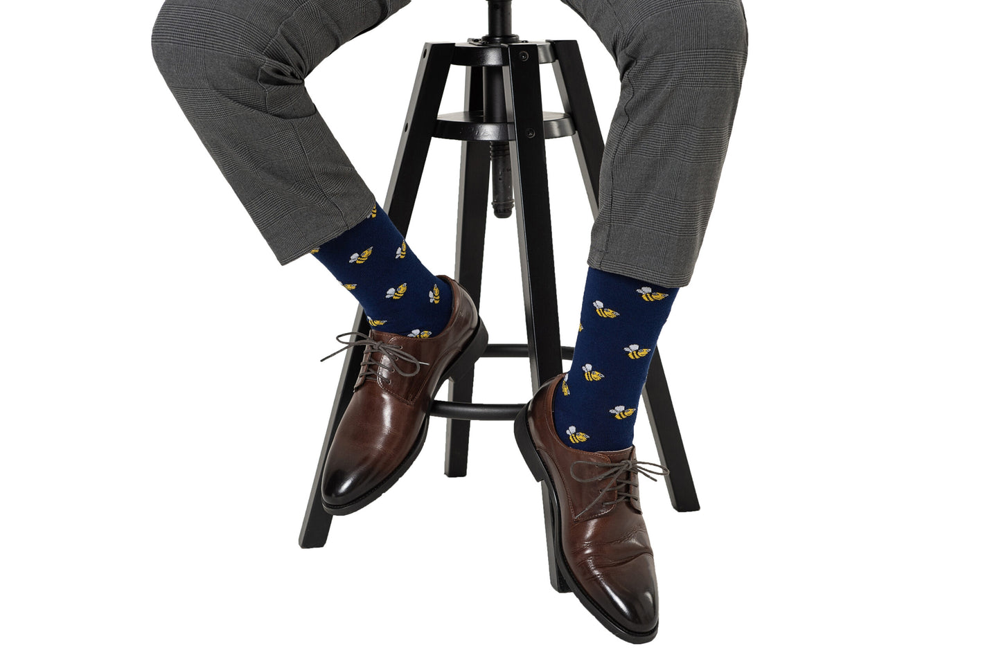 A person sits on a black stool, wearing gray plaid pants, dark brown dress shoes, and Bee Socks—navy blue socks featuring a yellow bee pattern.
