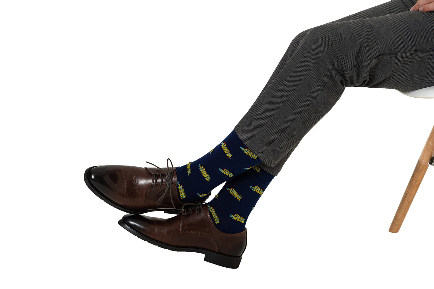A person wearing brown dress shoes, gray trousers, and navy blue Bus Driver Socks with yellow submarine patterns sits on a chair with legs crossed, exuding a sense of nostalgia.