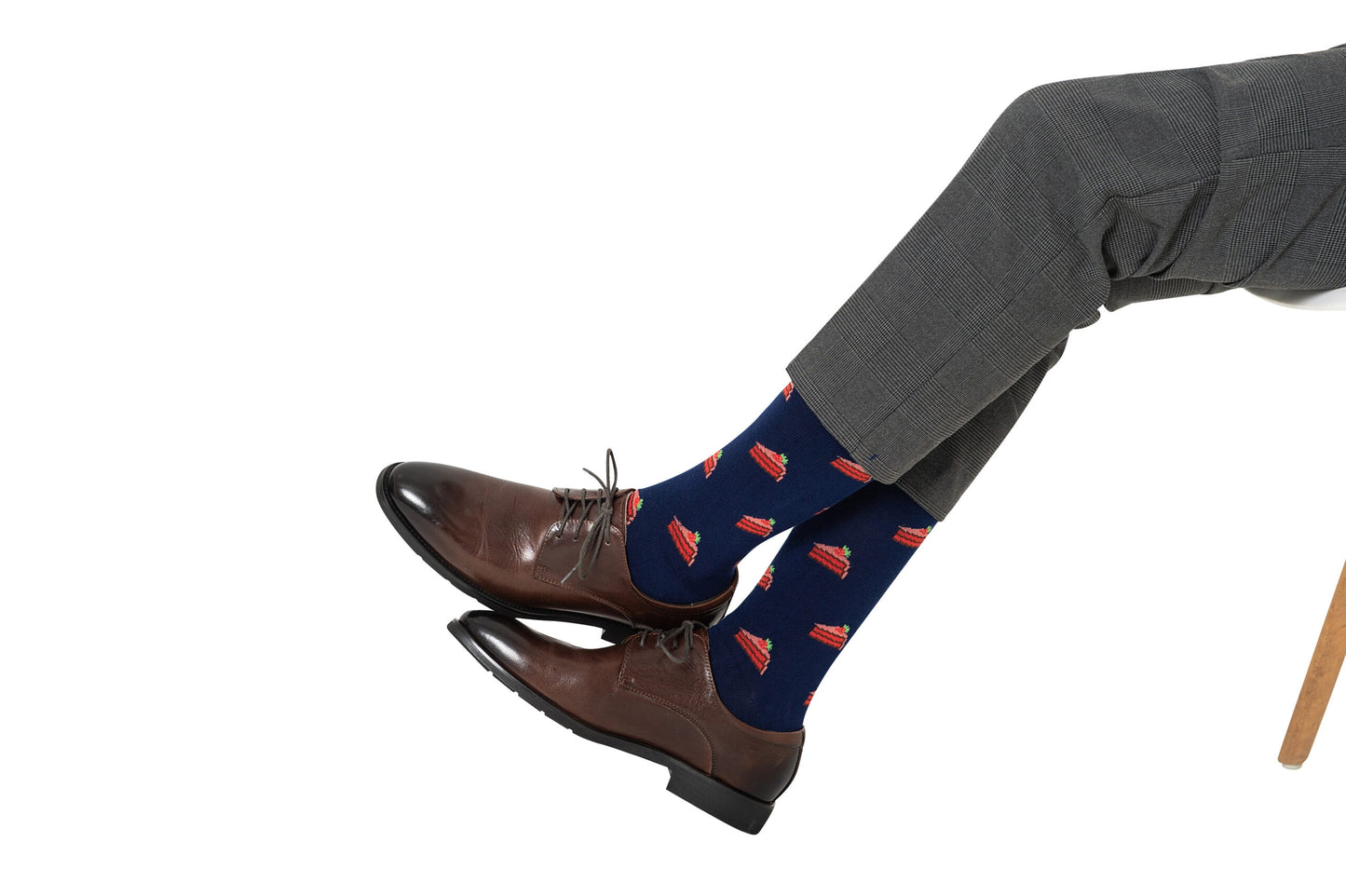 A person in gray dress pants, brown leather shoes, and Strawberry Cake Socks—a fun treat for dessert lovers—sits on a white chair.