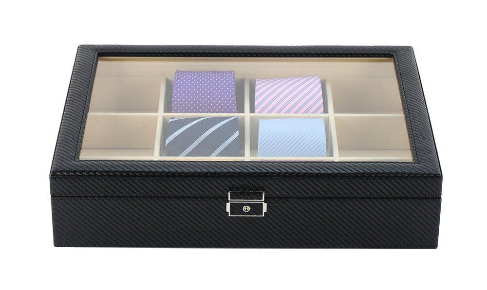 The Carbon Fibre Tie Box is a black display case with a glass lid and six compartments, providing stylish tie storage while showcasing four neatly folded neckties in assorted colors and patterns.
