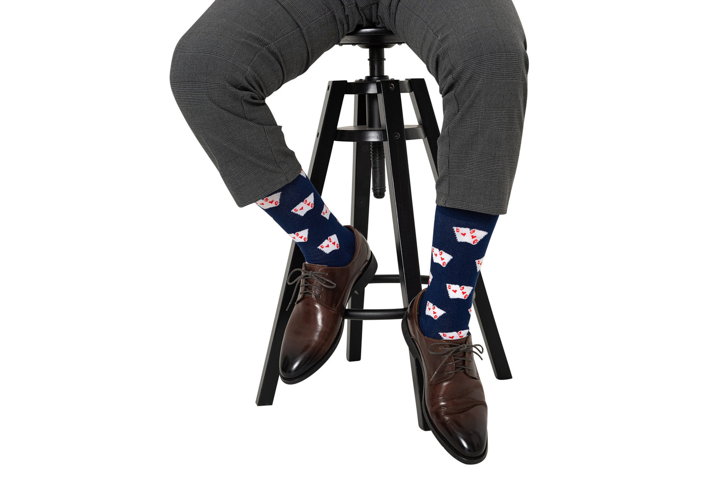 A person in gray checked pants, brown dress shoes, and Poker Socks featuring a playing card print sits cross-legged on a black stool—an ideal gift for poker enthusiasts.