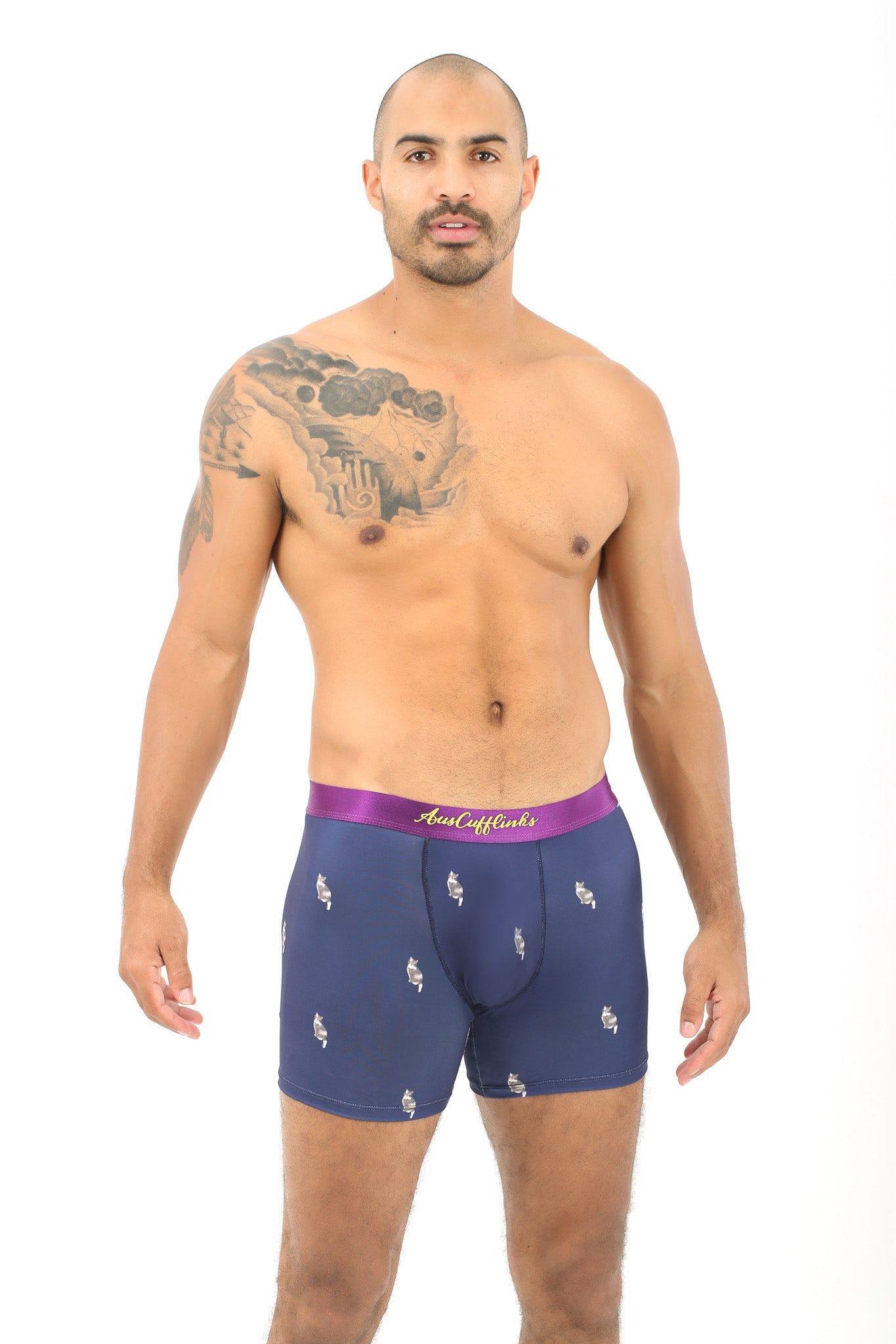 A man with a shaved head and tattoos on his arm is wearing Cat Underwear, navy blue boxer briefs featuring a purple waistband, embodying the snugness reminiscent of a cat's nine lives.
