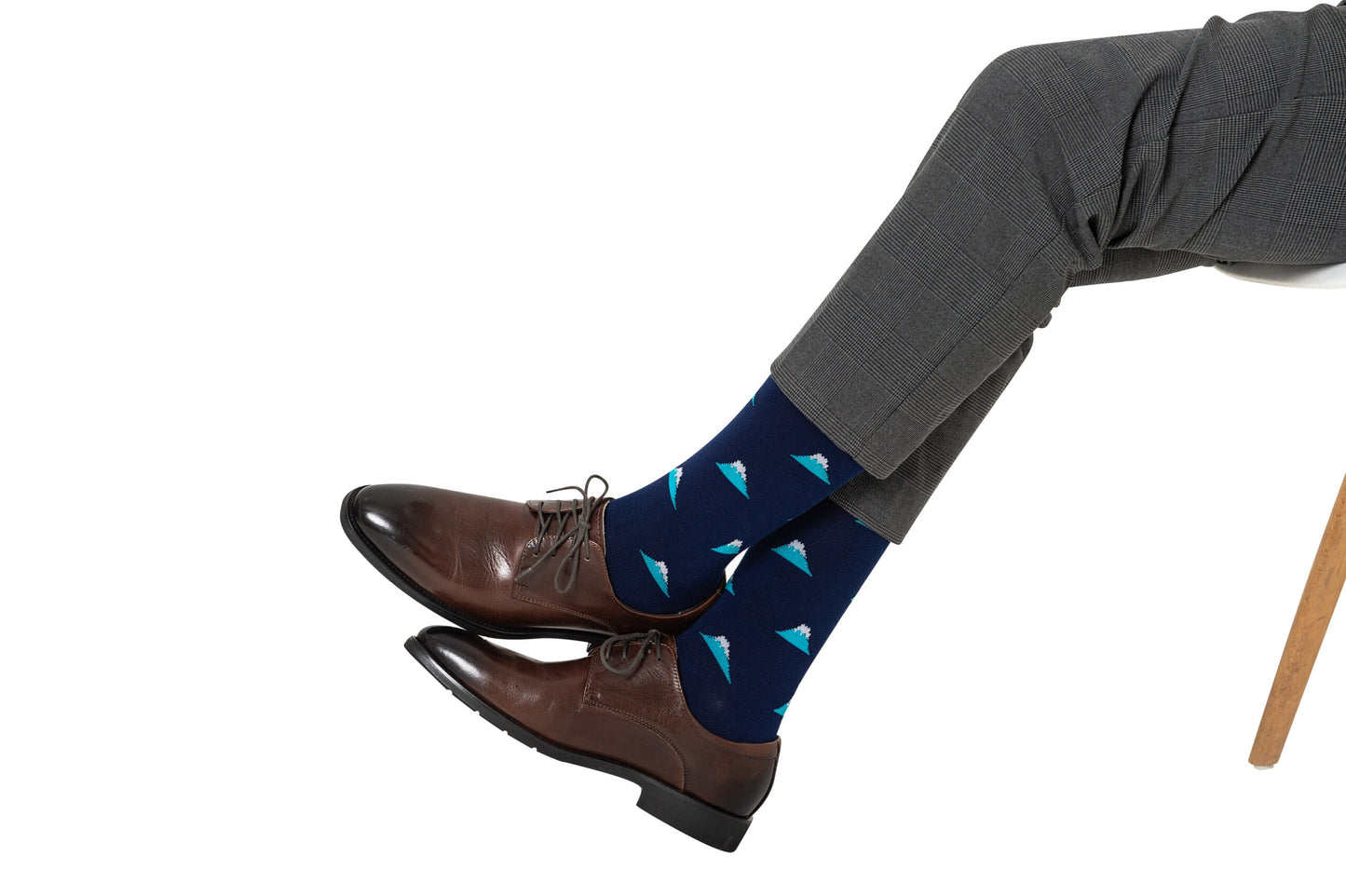 A person in gray pants and brown dress shoes with Mountain Socks, which feature a pattern of small white and blue sailboats, sits cross-legged on a white chair, showcasing their peak style.