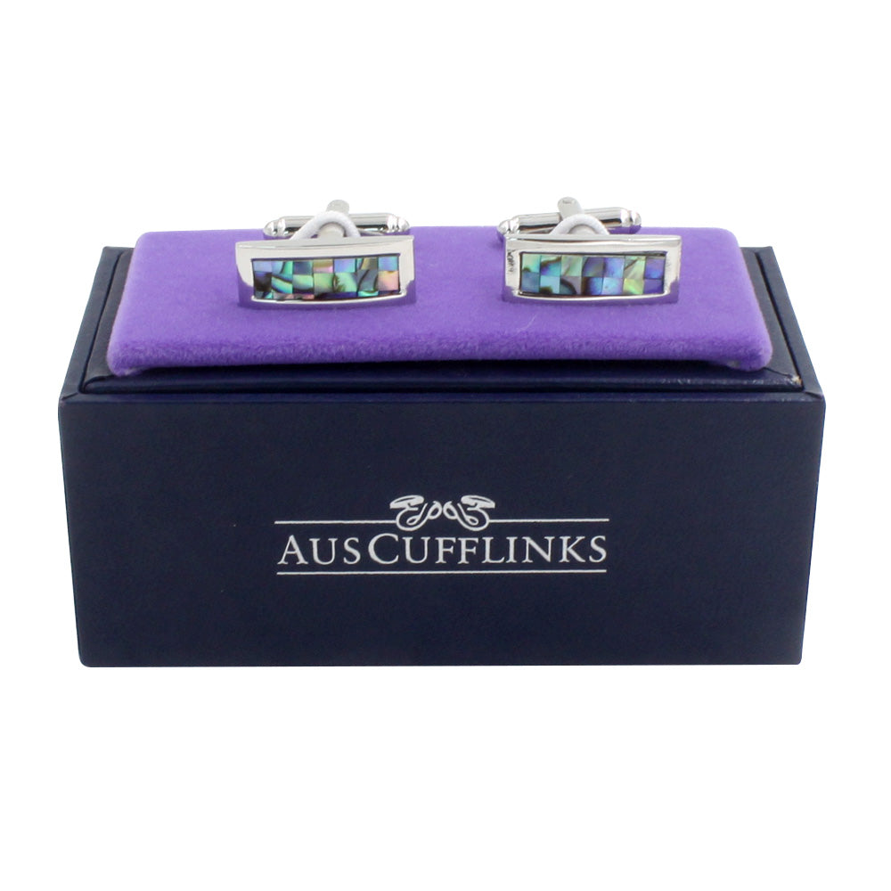 A pair of Mother of Pearl Stone Cufflinks, featuring rectangular metallic designs with abalone shell inlays that capture the ocean's depths and their lustrous allure, elegantly displayed on a purple cushion inside a black AUSCUFFLINKS branded box.
