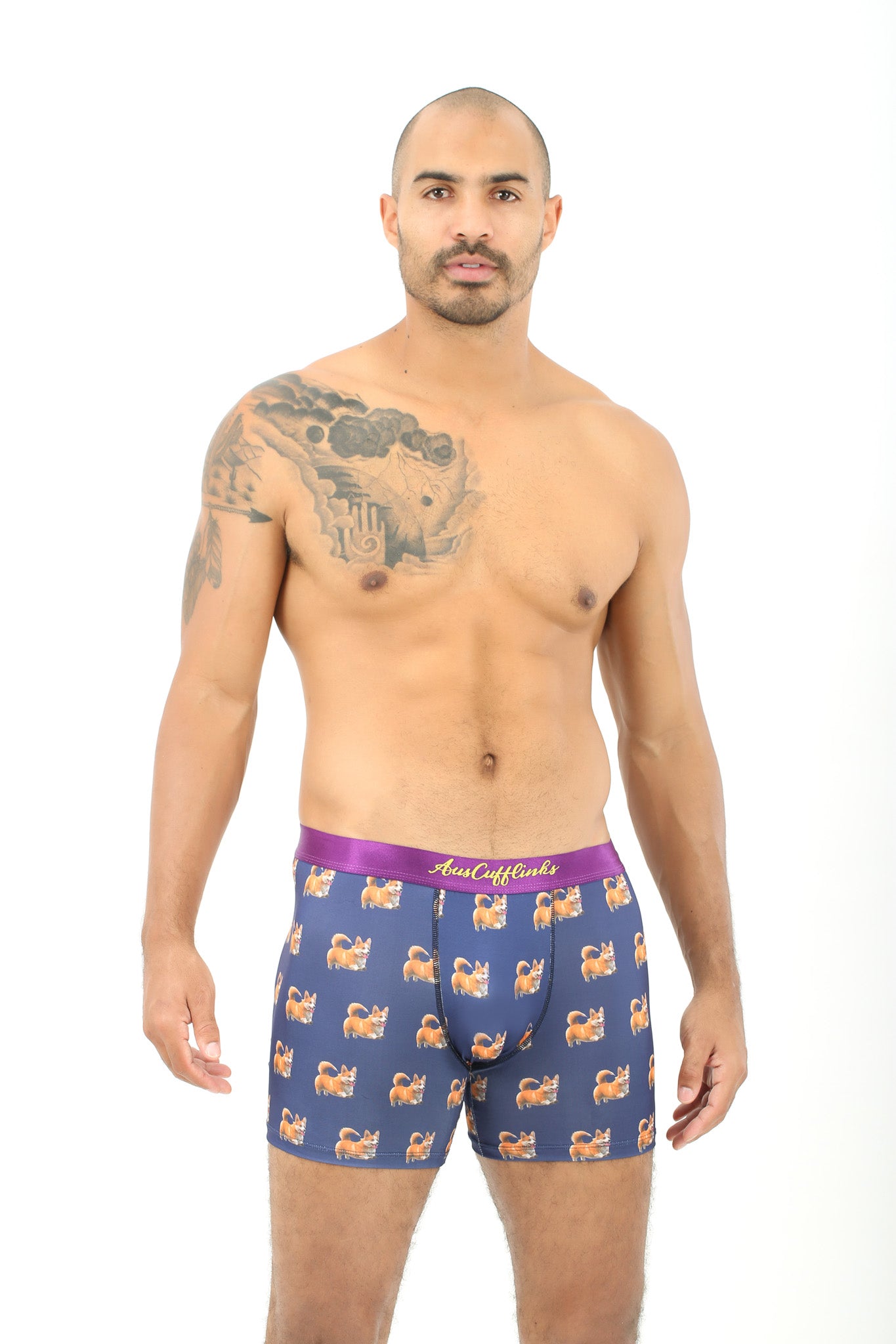 A shirtless man with a tattoo on his left shoulder wears blue Corgi Dog Underwear—fun boxers for dog lovers with a purple waistband—while standing against a plain white background.