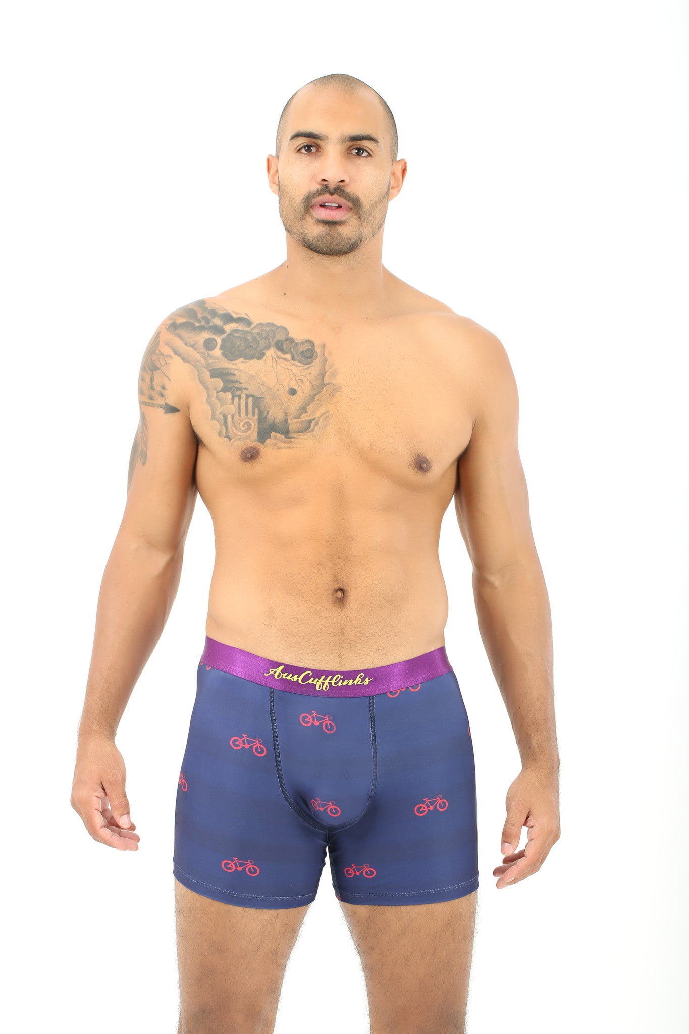 A man with a shaved head and chest tattoo models Cyclist Underwear – Fun Gift for Riders, featuring navy blue fabric with red bicycle patterns and a purple waistband, standing against a white background.