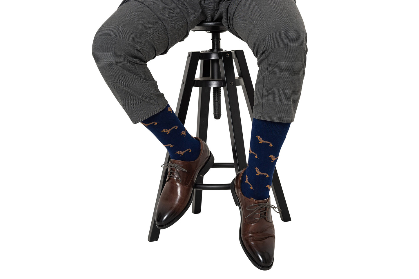 Wearing grey plaid pants and brown dress shoes, a person sits on a black stool, showing off Sausage Dog Socks—navy blue socks with a charming dachshund pattern.