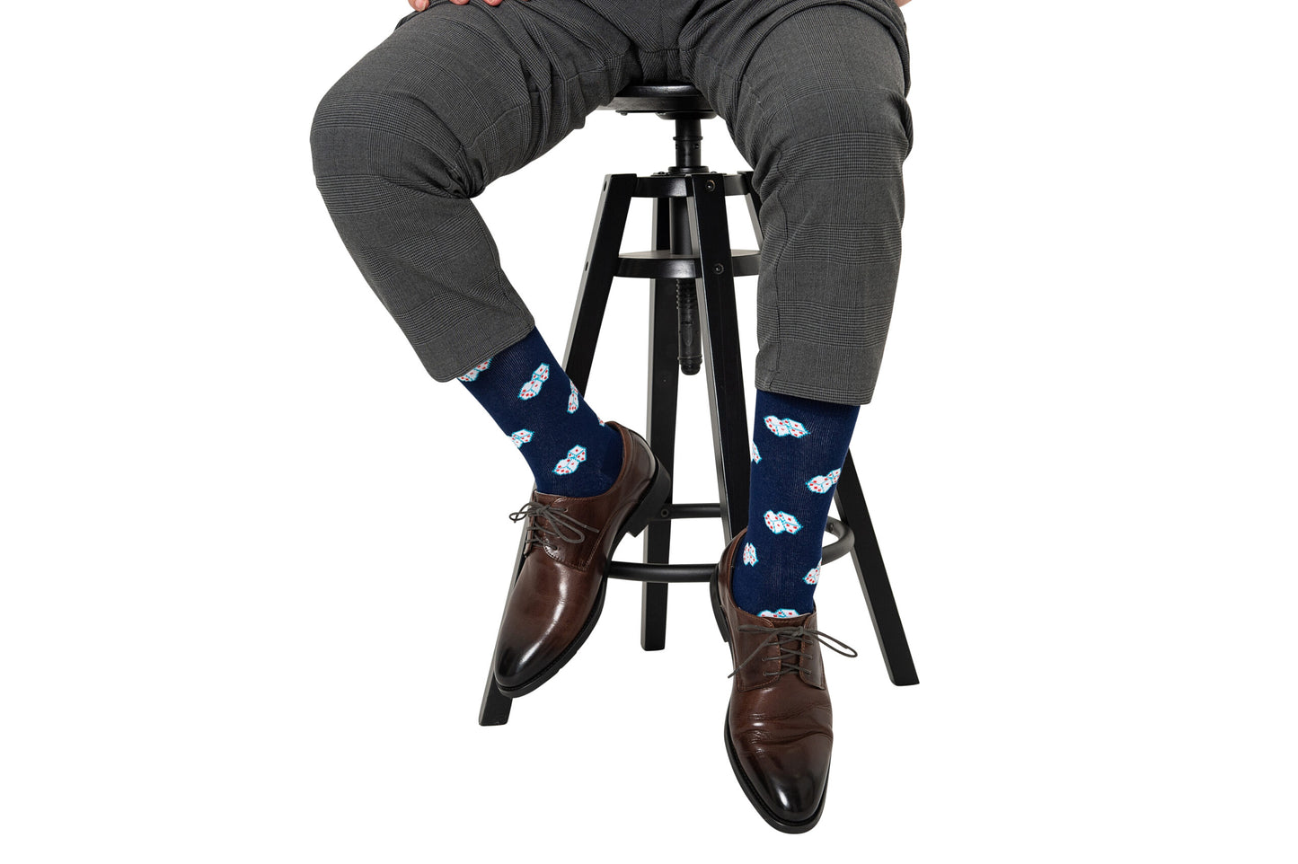 Man wearing gray dress pants, Dice Socks – Lucky Gift for Game Lovers, with blue patterns ideal for board gamers, and brown dress shoes, sits on a black stool against a white background.
