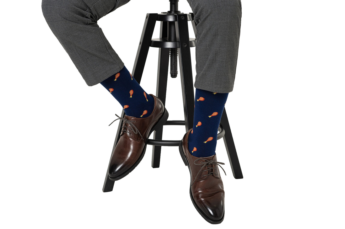 A person sweetly layered sits on a stool, wearing gray pants, Strawberry Cake Socks with a drumstick pattern, and brown leather shoes.