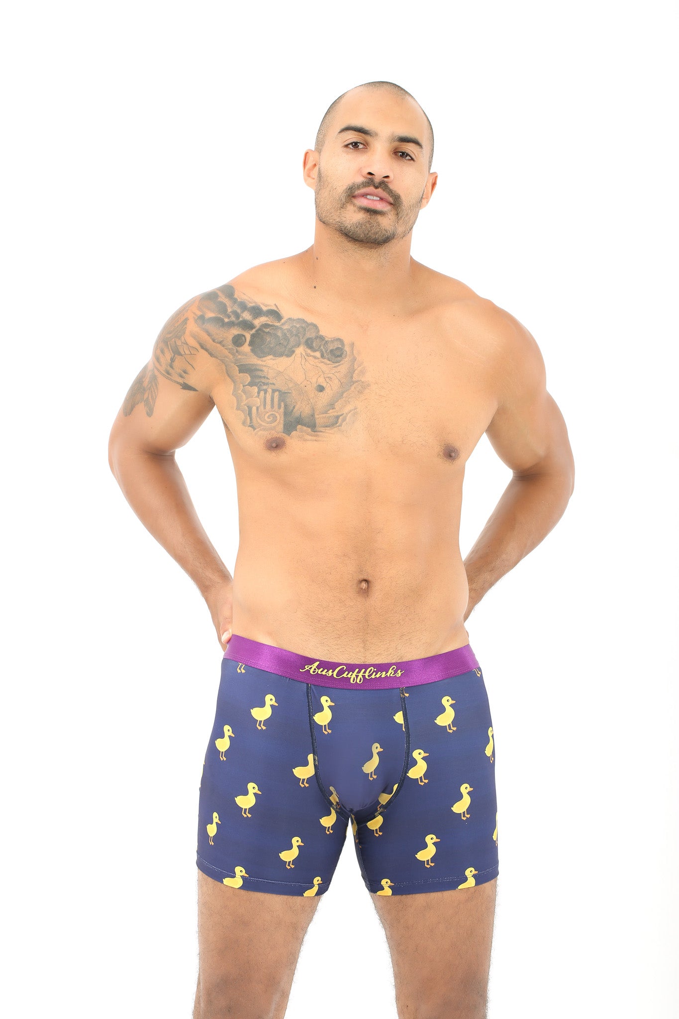 A shirtless man with a tattoo on his left shoulder faces forward, wearing AndCliffs Duck Underwear for Men—navy boxer briefs featuring a yellow duck pattern and purple waistband—ideal as a gift for animal and bird lovers.