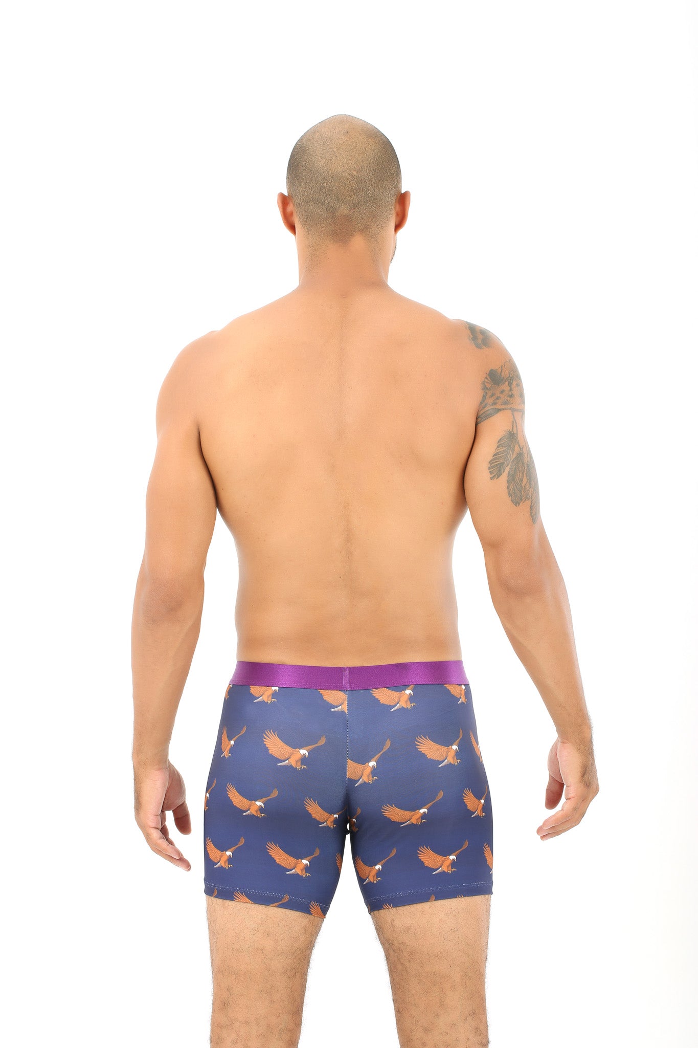 A man with a shaved head and a tattoo on his left arm stands with his back to the camera, wearing Eagle Pattern Underwear dark blue boxer briefs that evoke the coziness of open skies.