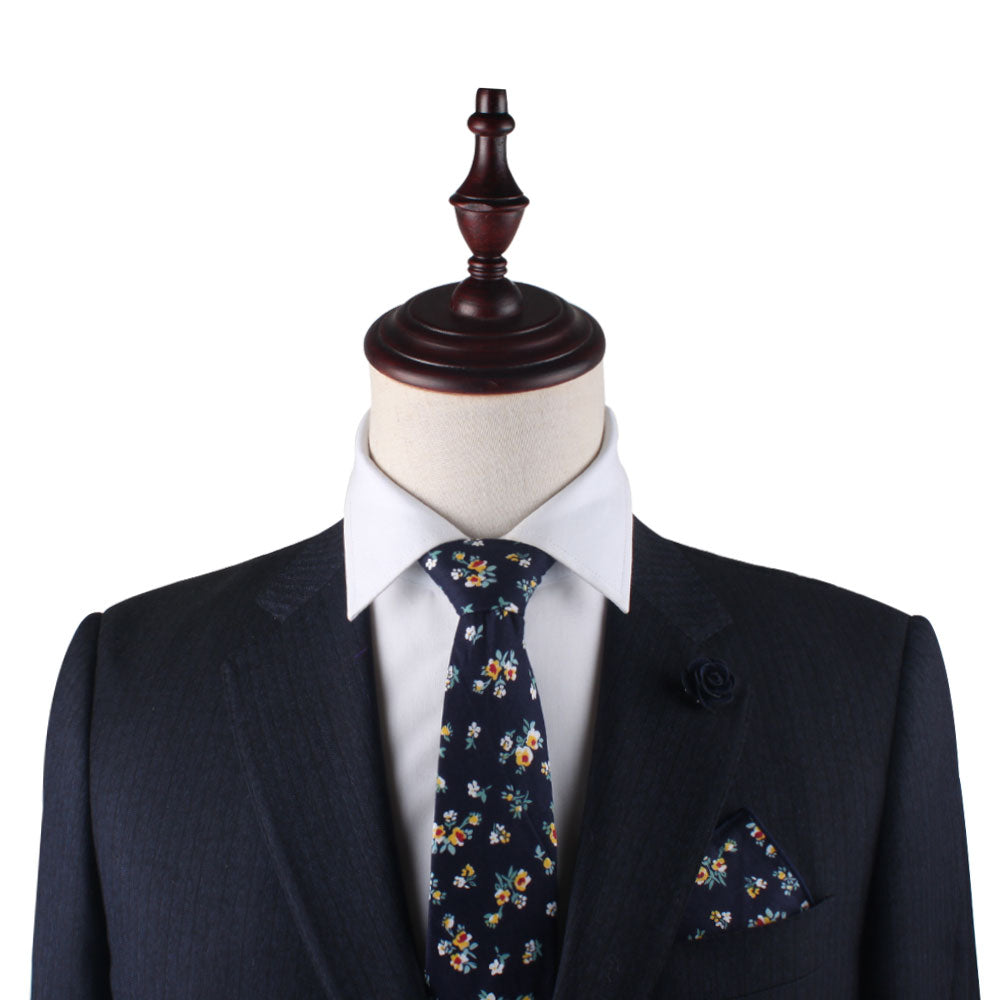 A mannequin elegantly dons a dark suit, complemented by a white shirt and the Floral Navy Yellow Skinny Cotton Tie and Pocket Square set, complete with a matching lapel pin.