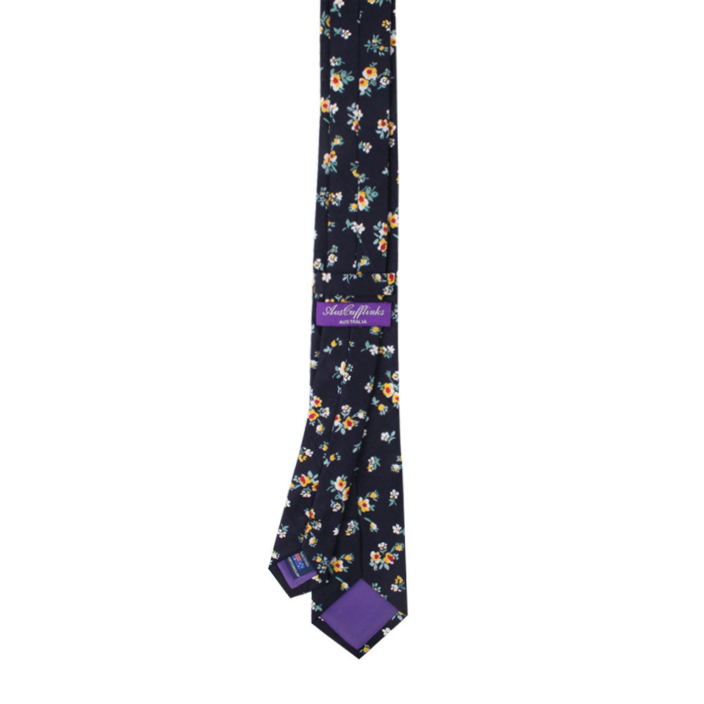 A navy skinny cotton necktie embellished with small yellow and white flowers, reminiscent of golden blossoms, is accompanied by a pocket square and features a purple label.