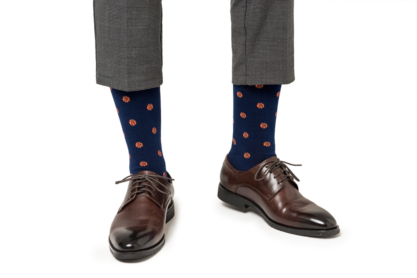 A person wears gray plaid pants, Basketball Socks for Men in navy with orange dots, and brown dress shoes, standing on a white background.