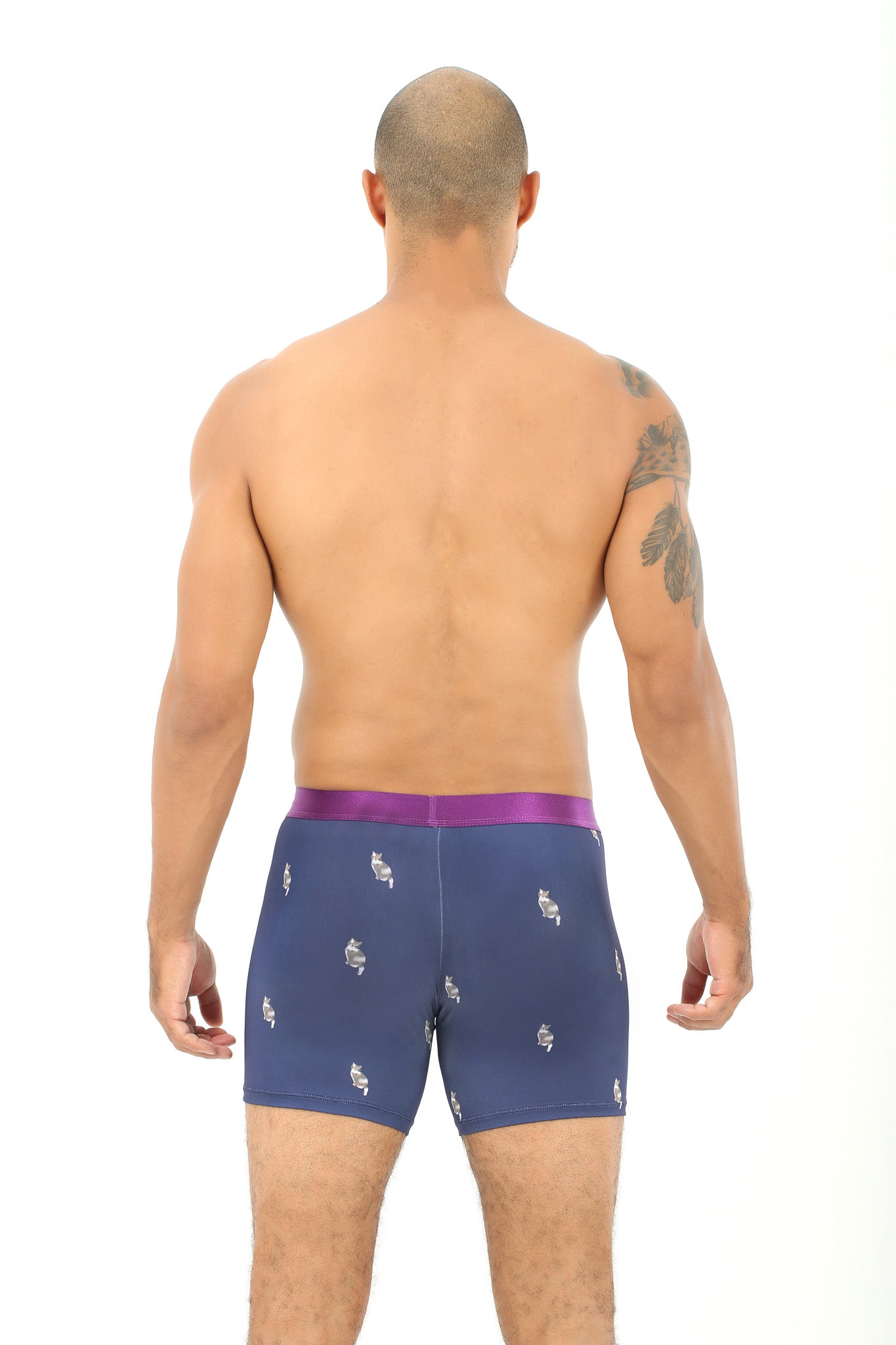 With their back to the camera, a person sporting a shoulder tattoo displays the snug fit of Cat Underwear, which are blue boxer briefs featuring a purple waistband, embodying the confidence of someone who feels like they have nine lives.