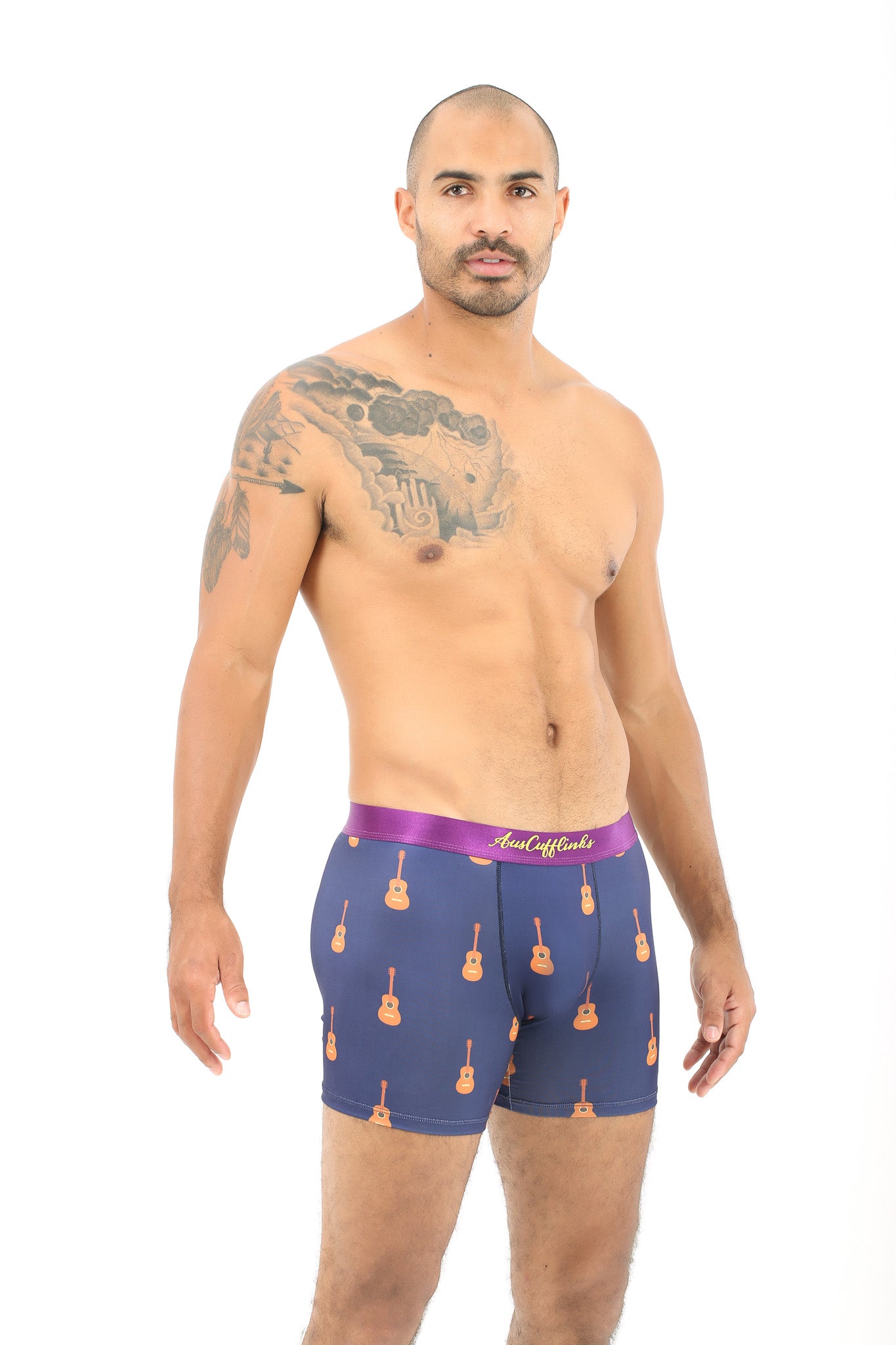 A man with a tattooed arm stands against a white background, wearing Guitar Underwear in blue, which captures the relaxed vibe of strumming chords.