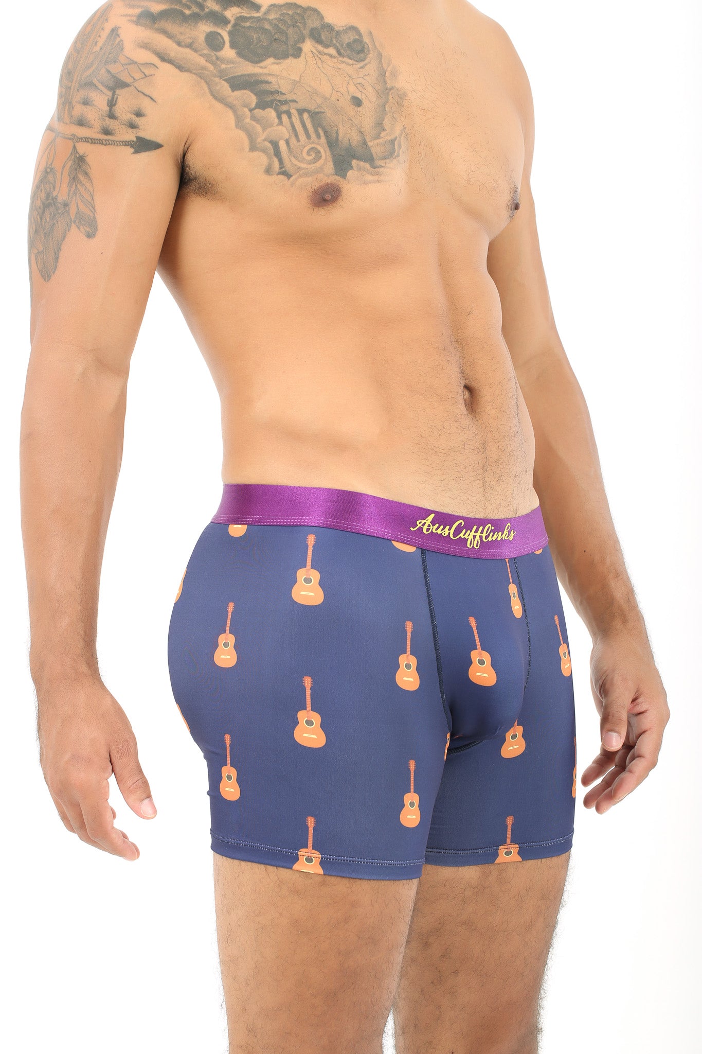 A man stands against a white background, mimicking guitar playing while wearing Guitar Underwear, which features a guitar pattern on blue boxer briefs with a purple waistband, effortlessly combining style and comfort.