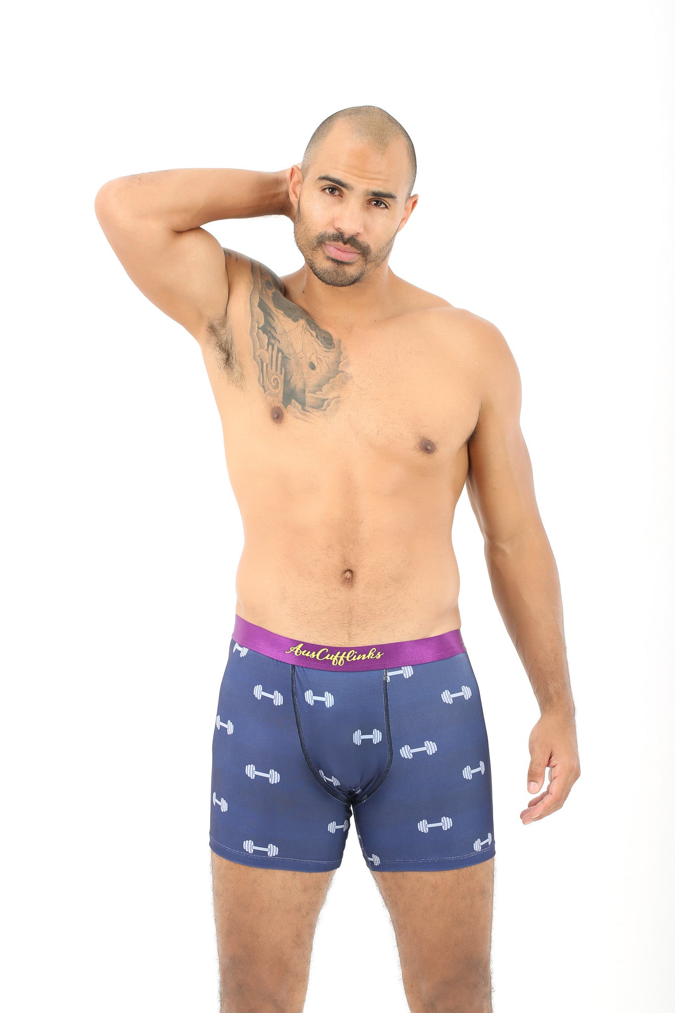 A man with a tattoo confidently poses in Gym Underwear, showcasing blue boxer shorts adorned with dumbbell patterns, bringing a playful game of style and comfort to life against a crisp white background.
