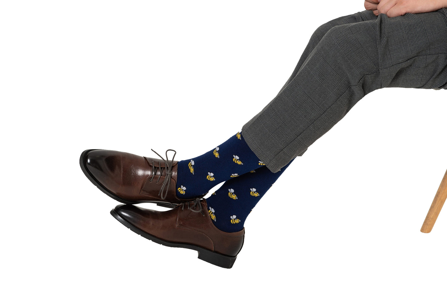 Someone wearing gray pants, brown dress shoes, and Bee Socks—blue socks featuring a yellow bee pattern—is seated with legs crossed.
