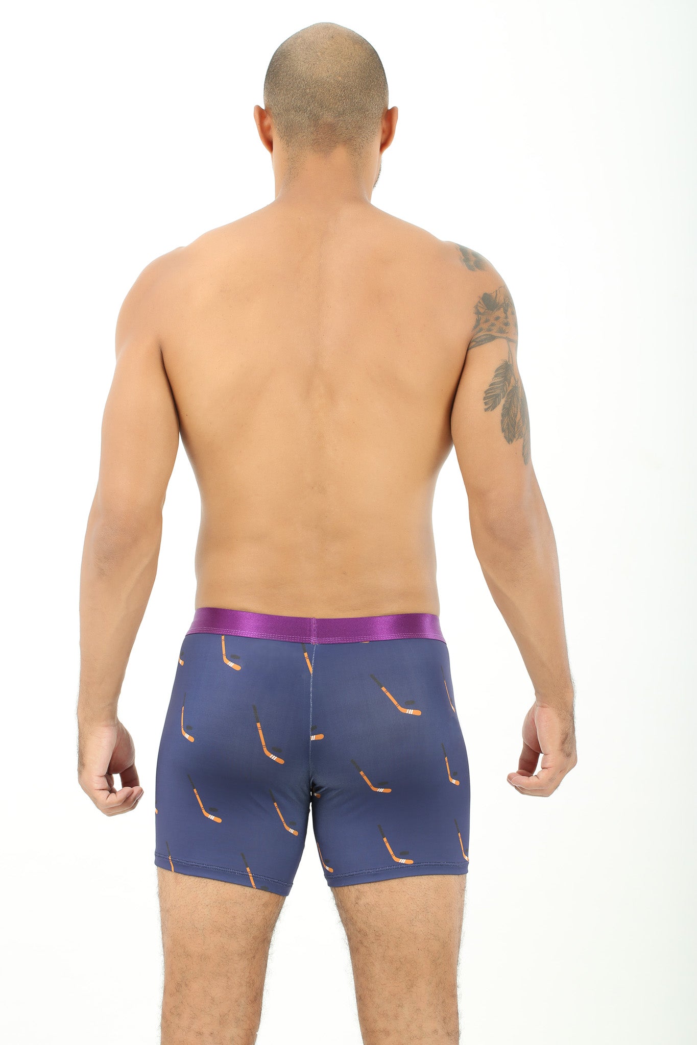 A man with a shaved head stands facing away, exuding chill vibes in Ice Hockey Stick Underwear featuring navy blue fabric with orange patterns and a purple waistband. His left shoulder boasts a tattoo, enhancing his look of effortless comfort.