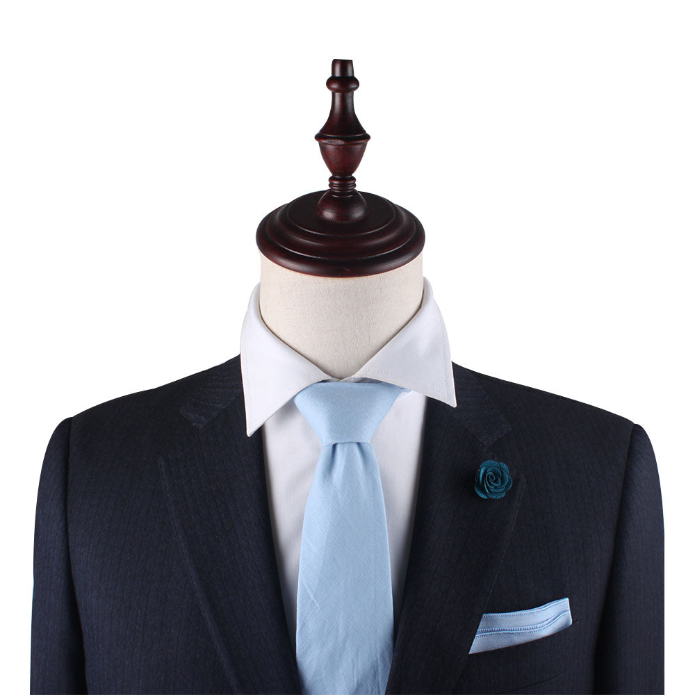 A suit displayed on a mannequin, featuring a dark blazer, light blue tie, white dress shirt, Light Blue Cotton Pocket Square, and a blue flower lapel pin exudes spring warmth with its seasonal blue accents.