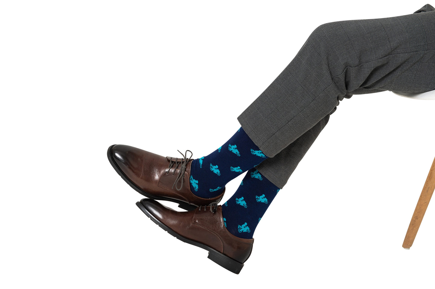 A person in gray dress pants and brown leather shoes sits with legs crossed, sporting Motorbike Socks—navy blue socks featuring light blue dinosaur patterns—making them a perfect biker gift for motorcycle lovers.