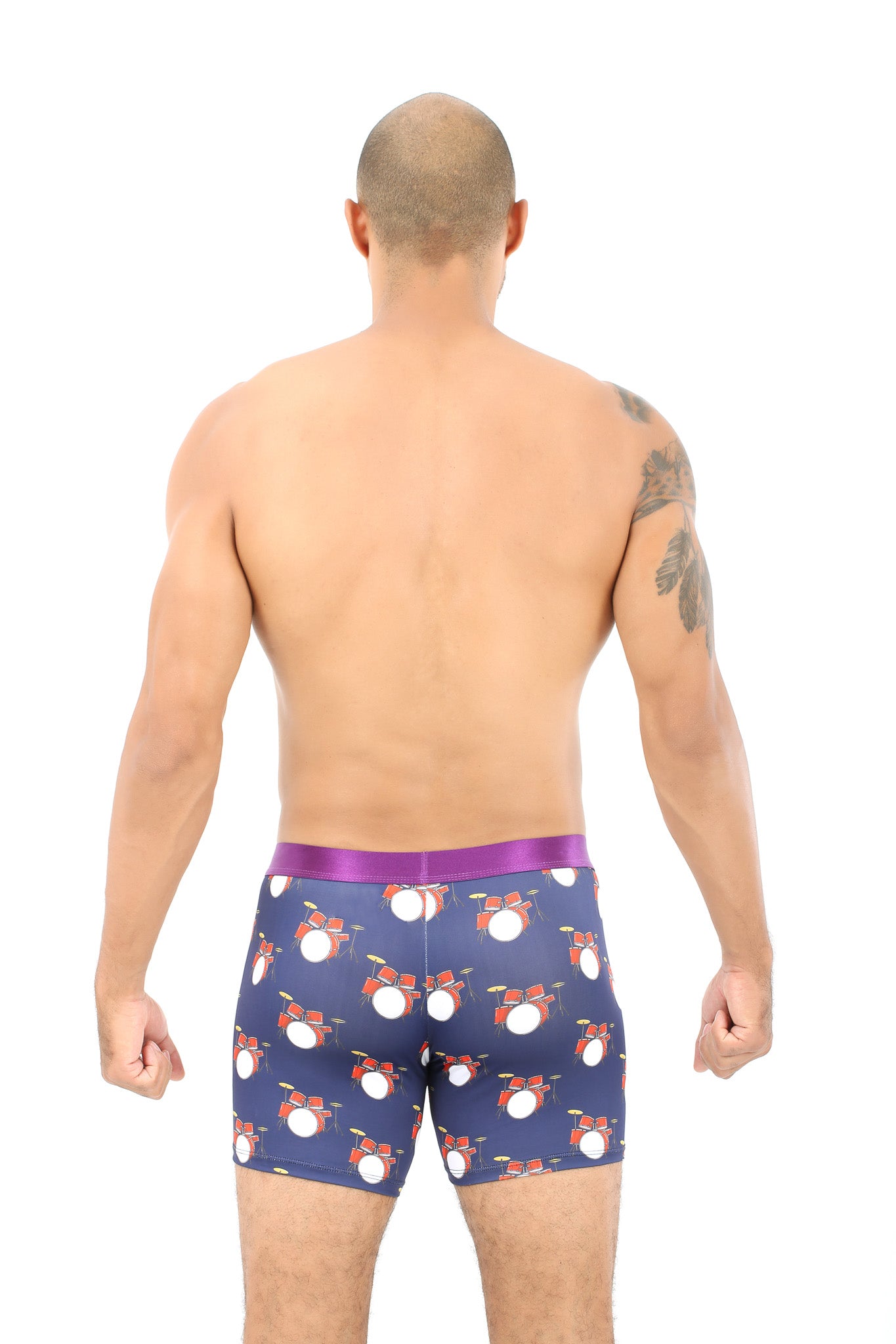 A man with a tattoo on his left arm stands with his back to the camera, wearing Drums Underwear in navy blue, showcasing a pink waistband and white and red drum patterns. The design captures the comfort rhythm of lively beats.